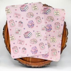 May include: Two pink notebooks with a repeating pattern of books, gifts, cats, armchairs, candles, and the text "Go away, I'm reading." The notebooks are on a wooden surface.