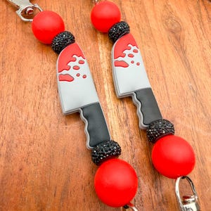 May include: Two keychains with a knife charm, red beads, and black rhinestone accents. The knife charms are gray with red blood splatter details and black handles. Each keychain has a silver clasp.