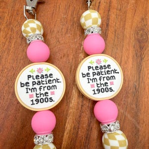 May include: Two beaded keychains with silver clasps. Each keychain features a pink bead, a rhinestone spacer, a gold and white checkered bead, and a round charm with the text "Please be patient. I'm from the 1900s."