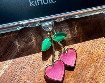 Puede incluir: Un amuleto dorado con dos cerezas de color rosa en forma de corazón y hojas verdes unidas a un lector electrónico Kindle.
