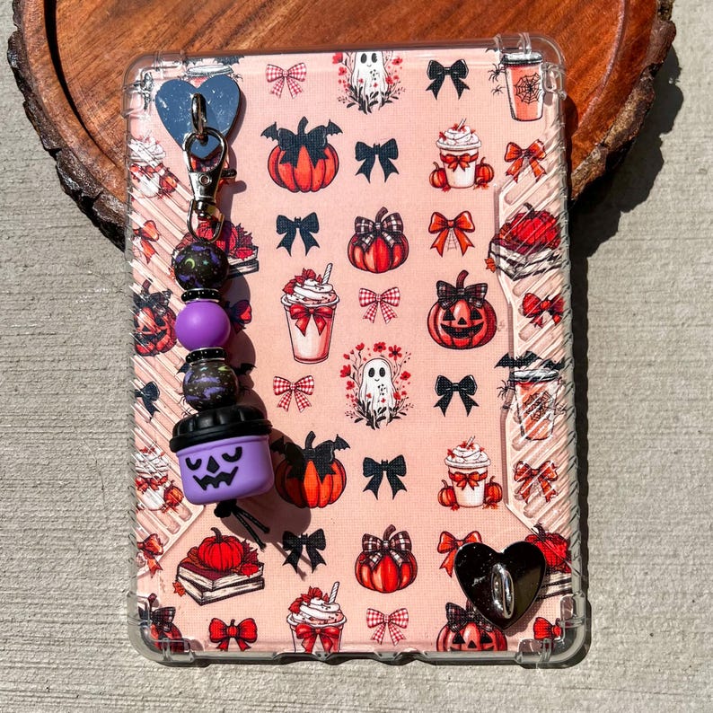 May include: A clear phone case with a pink background featuring pumpkins, bows, ghosts, and coffee cups. A charm with purple beads and a purple jack-o'-lantern is attached. A black heart-shaped button is on the case.