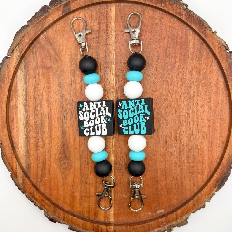 May include: Two beaded keychains with the text "ANTI SOCIAL BOOK CLUB" on black square beads. The keychains have black, white, and turquoise beads, and silver-colored clasps. The keychains are displayed on a wooden surface.