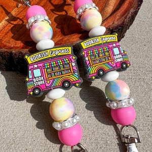 May include: Two beaded keychains with a pink and tie-dye color scheme. Each keychain features a pink bead, a tie-dye bead, and a charm shaped like a pink and yellow bookmobile with the text "BOOKISH EXPRESS BOOK STORE". The keychains have silver clasps.