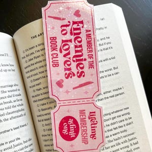 May include: A pink bookmark with the text "Enemies to Lovers Book Club" and "A Member of the Lifetime Membership". The bookmark has a ticket design with hearts and stars.