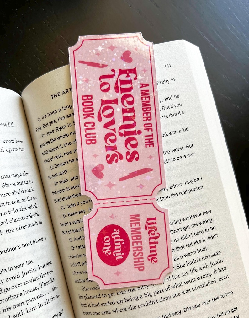 May include: A pink bookmark with the text "Enemies to Lovers Book Club" and "A Member of the Lifetime Membership". The bookmark has a ticket design with hearts and stars.