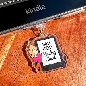 May include: A clear acrylic charm with the text "MOST LIKELY Reading Smut" on a tablet design. The charm features a pink heart pierced by an arrow and a flame design. The charm is attached to a Kindle.