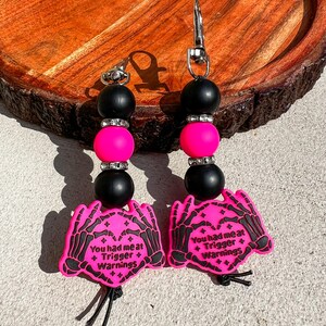 Op de afbeelding: Twee sleutelhangers met roze en zwarte kralen en een karabijnhaak. Elke sleutelhanger heeft een roze bedel met een skelet hand ontwerp en de tekst "You had me at + Trigger + Warnings."
