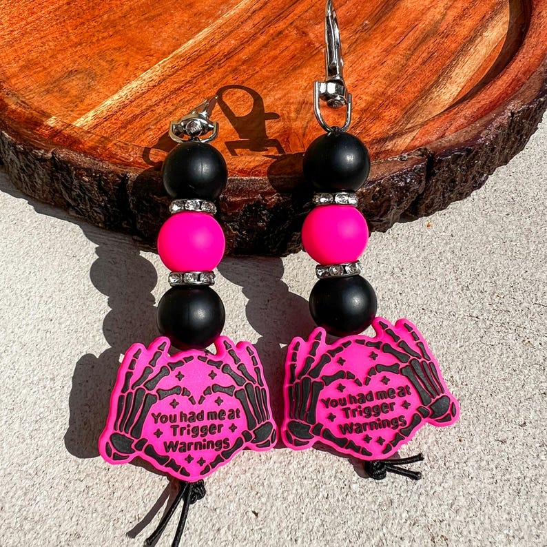 Op de afbeelding: Twee sleutelhangers met roze en zwarte kralen en een karabijnhaak. Elke sleutelhanger heeft een roze bedel met een skelet hand ontwerp en de tekst "You had me at + Trigger + Warnings."