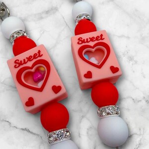 May include: Close-up of two pacifier clips with a Valentine's Day theme. Each clip features a pink rectangular charm with a heart-shaped cutout, the word "Sweet", and small red hearts. Red and white beads are also present.