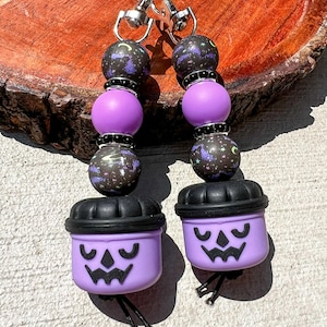 May include: A pair of Halloween-themed keychains. Each keychain features a purple pumpkin-shaped charm with a carved face, black beads, and purple and black speckled beads. The keychains are attached to silver-colored clasps.