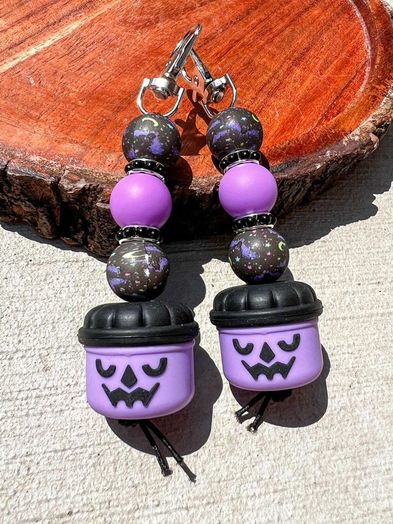 May include: A pair of Halloween-themed keychains. Each keychain features a purple pumpkin-shaped charm with a carved face, black beads, and purple and black speckled beads. The keychains are attached to silver-colored clasps.