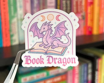 May include: A sticker featuring a pink dragon sitting on an open book, with the text "Book Dragon" in a decorative font. The dragon has purple wings and is set against a white background with celestial accents. The sticker is held by tweezers.