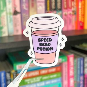 May include: A sticker featuring a coffee cup design with a pink and purple color scheme. The cup has the words "SPEED READ POTION" printed on the front. The sticker is surrounded by small star shapes.