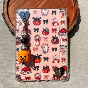 May include: A clear phone case with a pink Halloween-themed pattern featuring pumpkins, bows, ghosts, and coffee cups. A charm with a blue heart, beads, and an orange jack-o'-lantern is attached. The case is on a wooden surface.