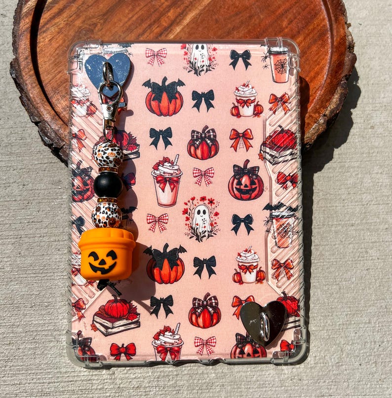 May include: A clear phone case with a pink Halloween-themed pattern featuring pumpkins, bows, ghosts, and coffee cups. A charm with a blue heart, beads, and an orange jack-o'-lantern is attached. The case is on a wooden surface.
