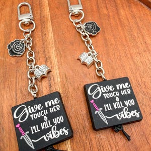 May include: Two black square keychains featuring the white text Give me TOUCH HER & I'LL KILL YOU vibes with a small pink and silver dagger graphic. Each keychain has a silver lobster clasp, a black rose charm, and an open book charm.
