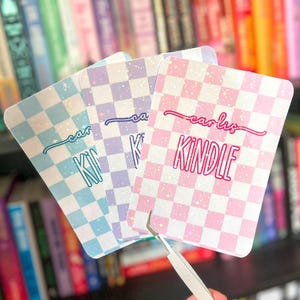 May include: Three rectangular Kindle covers with a checkered pattern in blue, purple, and pink. Each cover has the text "carls KINDLE" in a cursive font. The covers are held in front of a bookshelf.