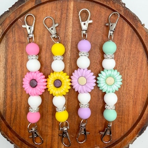 May include: Four beaded keychains with silver clasps. Each keychain features a different color scheme: pink, yellow, purple, and mint green. Each keychain has a flower charm, white beads, and rhinestone accents.