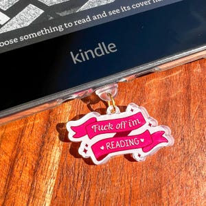 May include: A clear acrylic charm with a pink banner that reads "Fuck off i'm READING" in white text. The charm is attached to a Kindle device. The background is a wooden surface.