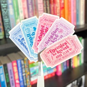 May include: A collection of colorful ticket-shaped stickers with text, held by tweezers. The stickers feature phrases like "Enemies to Lovers Book Club" and "Forbidden Love Club" in pink, blue, and purple hues. The background shows a bookshelf.