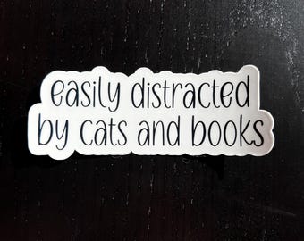 May include: White sticker with black text that reads "easily distracted by cats and books".