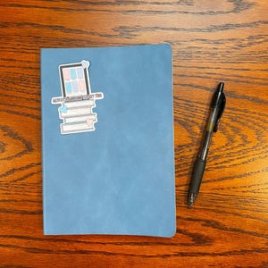 Może przedstawiać: Niebieski notes z naklejką z przodu, na kt&oacute;rej widnieje napis "Zawsze otaczaj się dobrymi wibracjami". Naklejka przedstawia stos książek z oknem na g&oacute;rze. Czarny długopis leży na drewnianej powierzchni obok notesu.