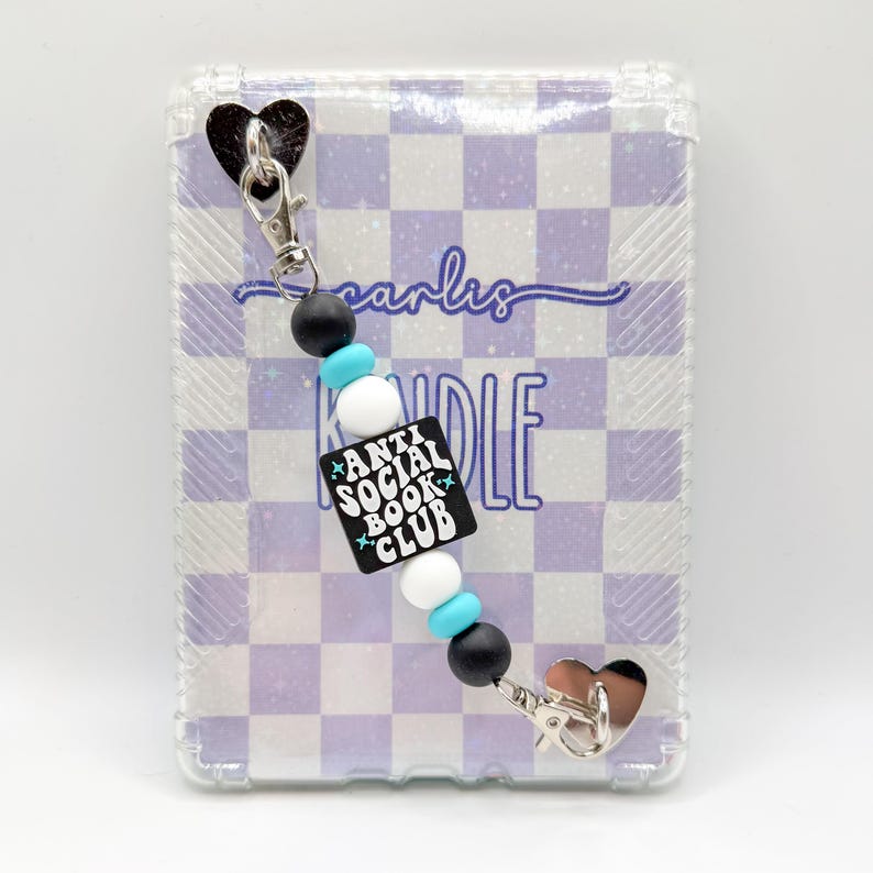 May include: A clear phone case with a purple and white checkered pattern. A beaded charm with the text "ANTI SOCIAL BOOK CLUB" is attached. The charm has black, white, and turquoise beads and silver-tone hardware.