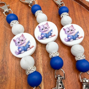 May include: Three lanyards featuring a central round bead with a graphic of a kitten reading a blue book. The lanyards are accented with alternating solid blue, white marble-patterned, and silver-toned rhinestone-encrusted spacer beads.