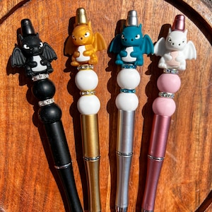 May include: Four dragon-themed pens with different colored bodies and dragon heads. The pens have a white bead and a clear bead with rhinestones.
