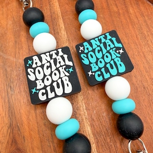 May include: Two keychains with black, white, and turquoise beads. Each keychain has a black square with the words "ANTI SOCIAL BOOK CLUB" in white and turquoise. The keychains have silver-colored clasps.