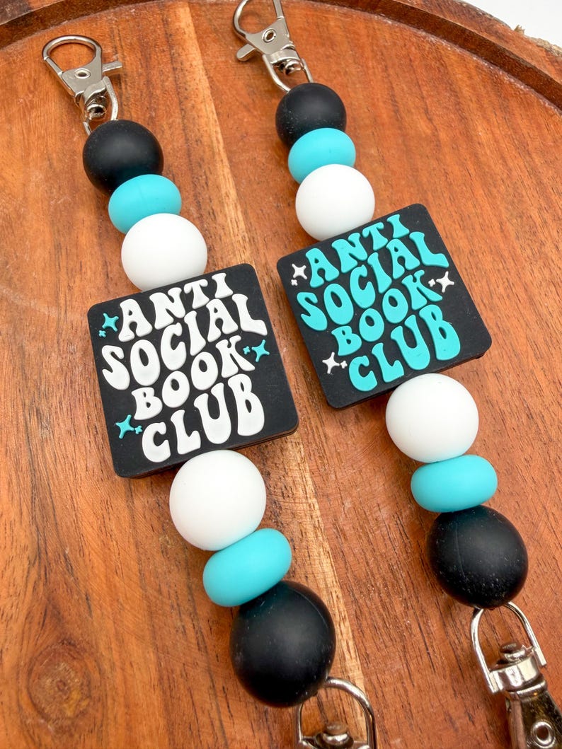 May include: Two keychains with black, white, and turquoise beads. Each keychain has a black square with the words "ANTI SOCIAL BOOK CLUB" in white and turquoise. The keychains have silver-colored clasps.