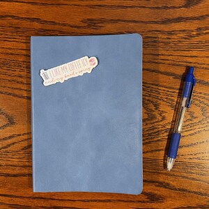 May include: A blue notebook with a sticker that says "I like my coffee icy and my books spicy". A blue pen is next to the notebook.
