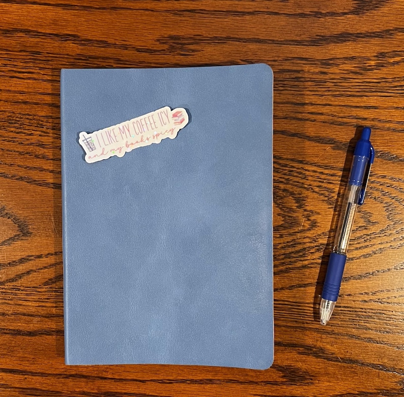 May include: A blue notebook with a sticker that says "I like my coffee icy and my books spicy". A blue pen is next to the notebook.