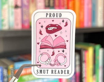May include: A pink sticker with the words "PROUD" and "SMUT READER". The sticker features illustrations of lips, a book, handcuffs, hearts, stars, and chili peppers. The sticker is held by a pair of tweezers.
