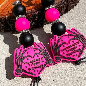 Op de afbeelding: Een kralen lanyard met zwarte en felroze kralen, met strass accenten. Twee felroze handvormige bedels met zwarte skeletontwerpen en de tekst "You had me at Trigger Warnings" zijn bevestigd.