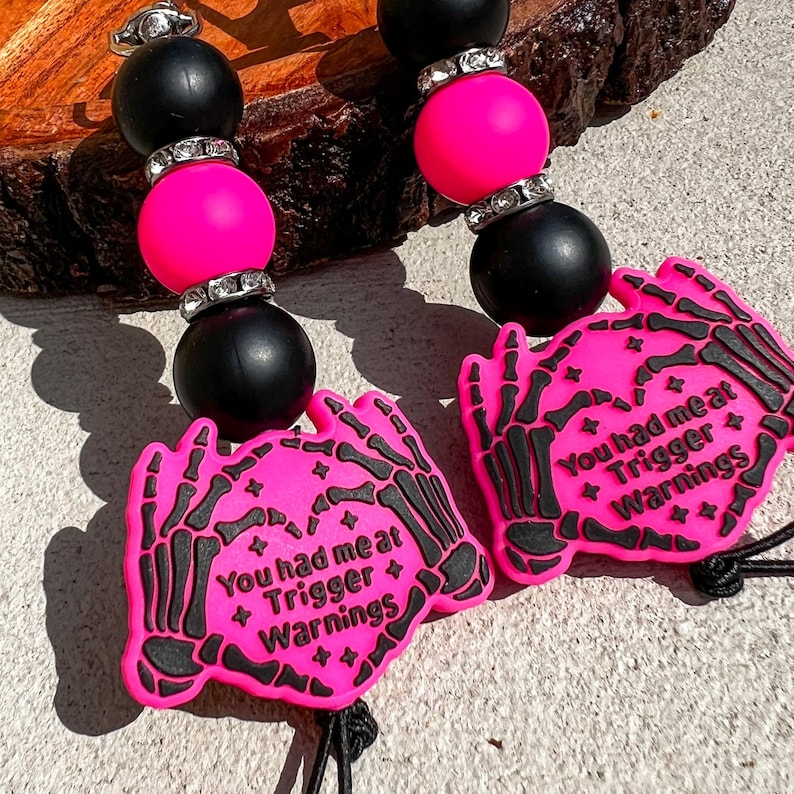 Op de afbeelding: Een kralen lanyard met zwarte en felroze kralen, met strass accenten. Twee felroze handvormige bedels met zwarte skeletontwerpen en de tekst "You had me at Trigger Warnings" zijn bevestigd.
