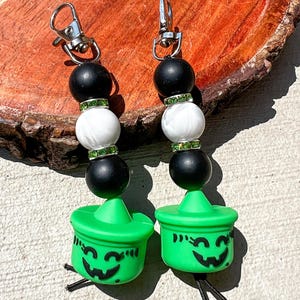May include: Two Halloween-themed keychains. Each keychain features a silver clasp, black and white beads, and a green jack-o'-lantern charm with a smiling face. The charms have black string tassels.