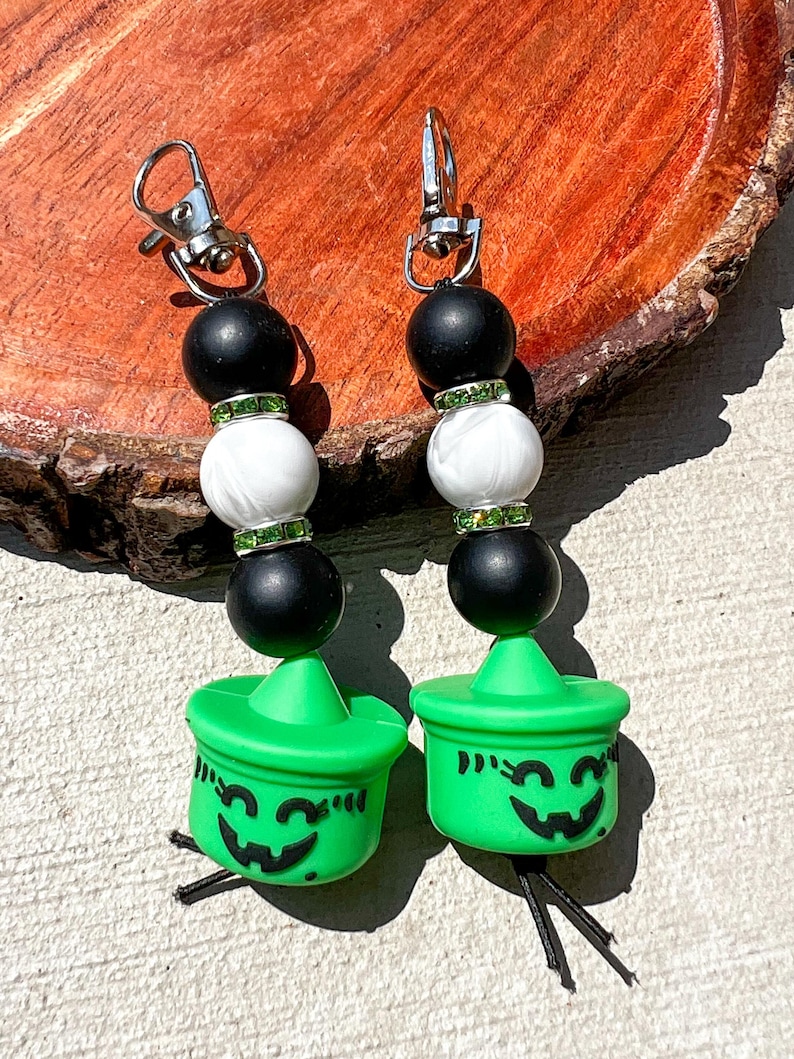 May include: Two Halloween-themed keychains. Each keychain features a silver clasp, black and white beads, and a green jack-o'-lantern charm with a smiling face. The charms have black string tassels.