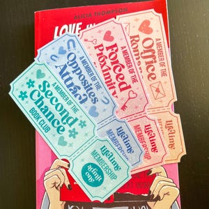 May include: A red book with the title "Love in" and decorative tickets. The tickets feature phrases such as "Forced Proximity" and "Opposites Attract." The tickets are light blue and pink with heart and key designs.