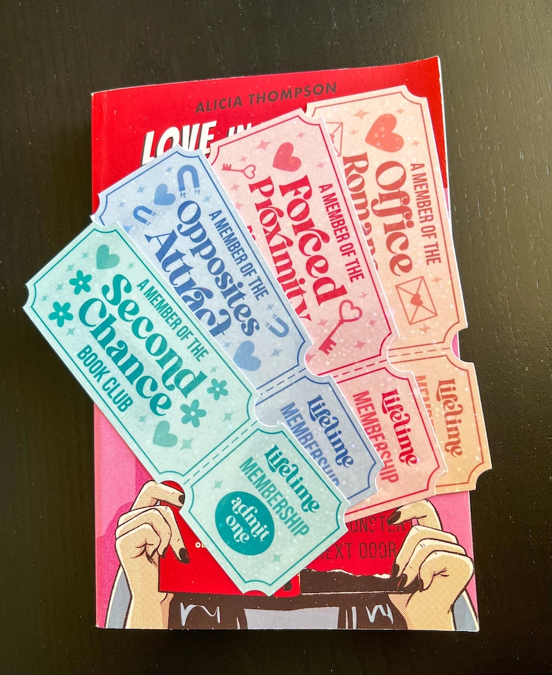 May include: A red book with the title "Love in" and decorative tickets. The tickets feature phrases such as "Forced Proximity" and "Opposites Attract." The tickets are light blue and pink with heart and key designs.
