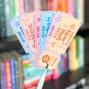 May include: A selection of pastel-coloured book club membership tickets. Each ticket displays a different design and text, including "Triangle Book Club" and "Slow Burn Book Club." The tickets are held by tweezers, highlighting their detailed designs and soft colour scheme.