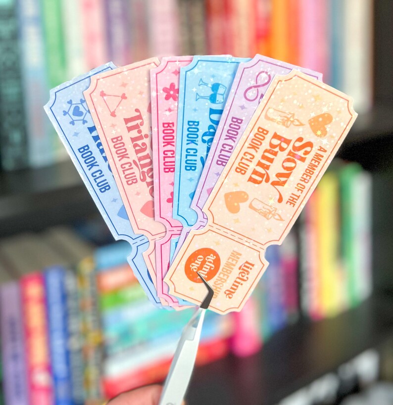 May include: A selection of pastel-coloured book club membership tickets. Each ticket displays a different design and text, including "Triangle Book Club" and "Slow Burn Book Club." The tickets are held by tweezers, highlighting their detailed designs and soft colour scheme.