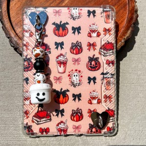 May include: A clear phone case with a pink background featuring pumpkin, bow, ghost, and whipped cream designs. A beaded charm with a white coffee cup, smiley face, and decorative beads is attached to the case.