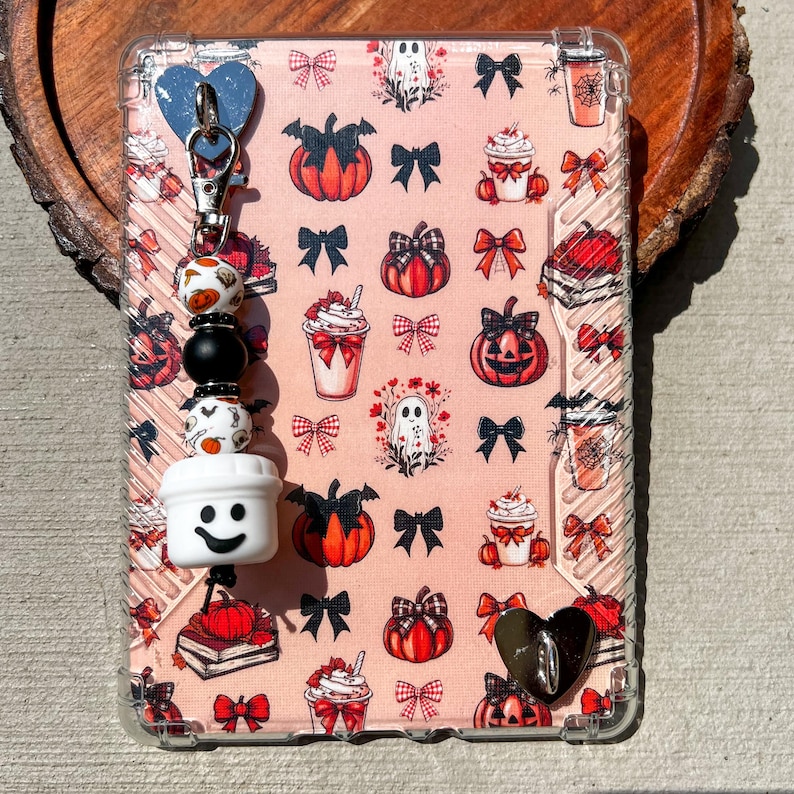 May include: A clear phone case with a pink background featuring pumpkin, bow, ghost, and whipped cream designs. A beaded charm with a white coffee cup, smiley face, and decorative beads is attached to the case.