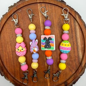 May include: Four Easter-themed keychains with colorful beads and charms. Each keychain features a different design: a chick in an egg, a bunny gnome, a Happy Easter sign with a bunny, and a decorated Easter egg. The keychains have silver-tone clasps.