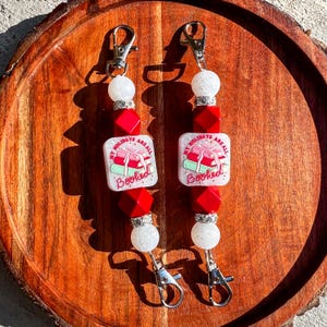 May include: Two keychains with silver clasps, white and red beads, and square charms. The charms have a stack of books and a gift with the text "My Holidays are all Booked". The keychains are on a wooden tray.