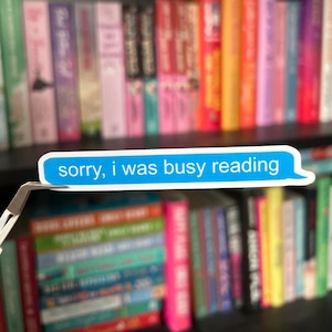 May include: A blue and white sticker with the text "sorry, i was busy reading" on a bookshelf with colorful books.