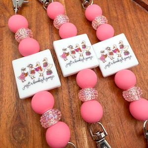 May include: Three beaded lanyards featuring pink silicone spheres, sparkling pink rhinestone beads, and a white square charm. The charm displays a graphic of four geese reading books with the text "just a bookish goose" in black script.