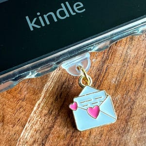 May include: A gold charm in the shape of a white envelope with pink hearts attached to a clear plastic book holder. The book holder is attached to a black Kindle e-reader.