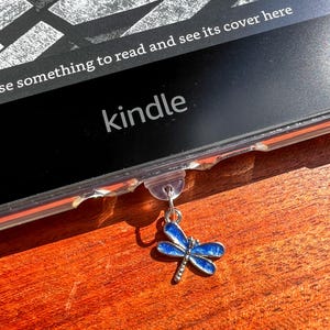 May include: A blue dragonfly charm with silver accents hangs from a clear Kindle case. The Kindle is black with the word "kindle" in white. The text "see something to read and see its cover here" is also visible.